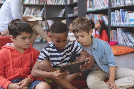 Overhead view of schoolboys using a digital tablet while they are sitting on cushions in a library at school with their teacher reading a story to their classmates in background