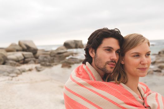 Side view of Caucasian couple wrapped in blanket standing at beach om a sunny day. They are hugging each other.