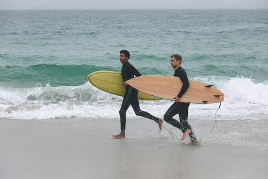 Side view of young multi-ethnic male friends running on beach while holding surfboards