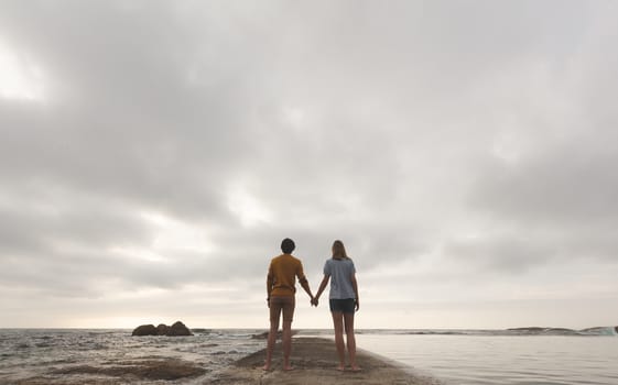 Rear view of young Caucasian couple standing hand in hand on rock near beach 