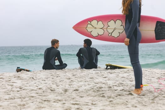 Rear view of multi ethnic male friends interacting with each other while woman standing with surfboard on beach 