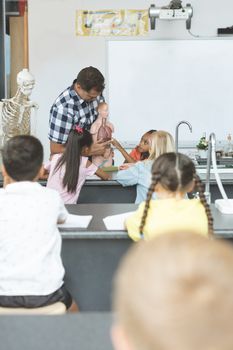Front view of a teacher holding a dummy skeleton while a schoolgirl touching his lungs in classroom at school with scholl kids sitting on their chair in foreground