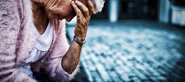 Side view of depressed senior woman sitting on bench with head in hand against nursing home