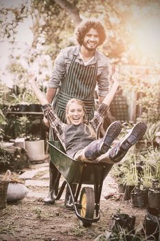 Portrait of cheerful man giving wheelbarrow ride to female gardener outside greenhouse