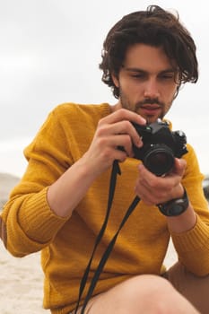 Front view of young Caucasian man using digital camera sitting on the beach 