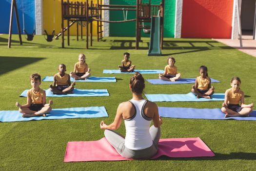 Rear view of caucasian trainer teaching yoga to students on yoga mat in school playground at schoolyard