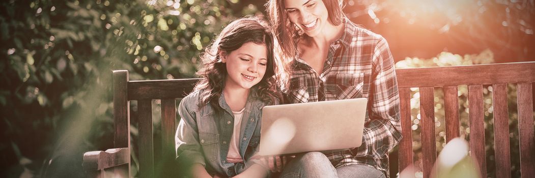 Smiling mother and daughter using laptop while sitting on wooden bench at backyard