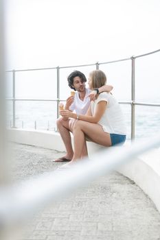 Front view of young Caucasian couple siting and interacting at promenade while having ice cream cone. They are smiling