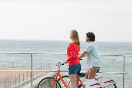 Side view of young Caucasian couple interacting with each other while holding bicycle at promenade near beach