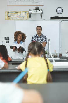 Front view of an African ethnicity schoolboy next to his teacher and holding a book  while looking at his classmates