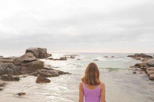 Rear view of young Caucasian blonde woman standing at beach. She is looking the sea 