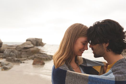 Side view of young Caucasian couple wrapped in blanket kissing each other standing at beach  
