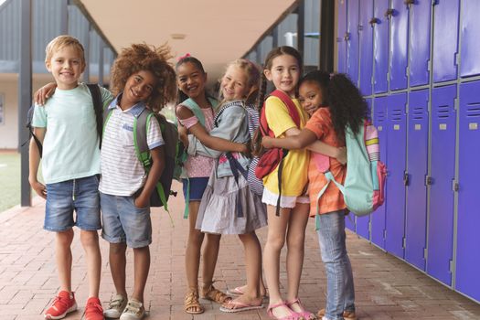 Front view of happy school kids standing in corridor and holding together in arms