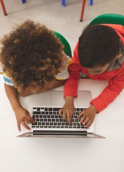 Overhead view of two mixed-race school boys taping on the keyboard of their laptop in classroom at school