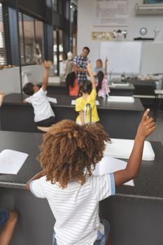 Rear view of school kids raising hand in classroom at school with teacher in background
