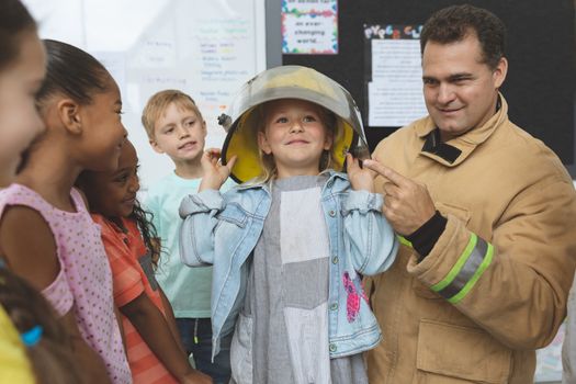 Front view of a Caucasian firefighter teaching to school kids about fire security while one of them has a fire helmet over her head