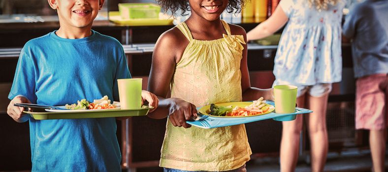 Portrait of happy school children holding food tray in canteen against classmates