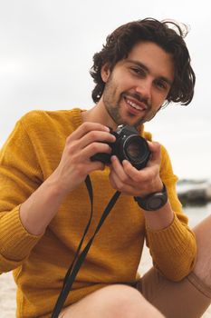 Front view of young Caucasian man using digital camera sitting on the beach. He is smiling and looking at camera