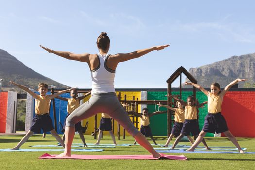 rear view of caucasian trainer teaching yoga to students on yoga mat in school playground at schoolyard