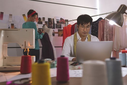 Male fashion designer working on laptop while female fashion designer dressing mannequin in design studio 