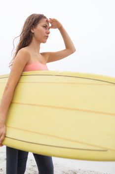 Rear view young Caucasian female surfer standing with her surfboard on the beach. She is looking away