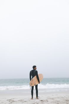 Rear view of thoughtful young mixed-race male surfer holding surfboard on the beach. He is looking away