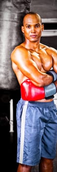 Portrait of confident male boxer standing with arms crossed by punching bag in fitness studio