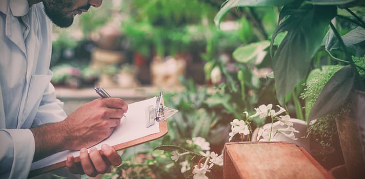 Side view of male scientist writing in clipboard while examining plants at greenhouse