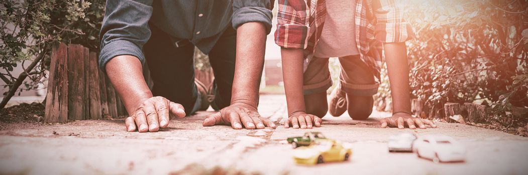 Grandson and grandfather playing with toy cars while kneeling on pavement in yard