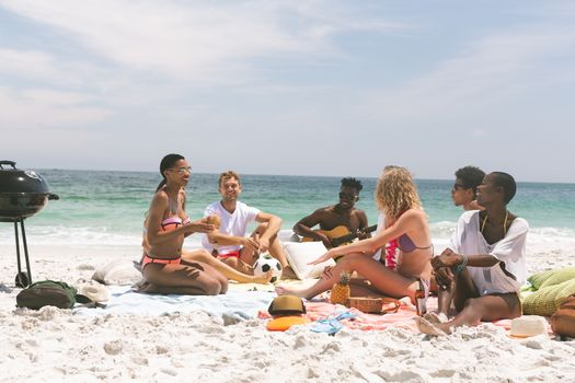 front view of multi ethnic group of friends relaxing and sitting on beach while interacting with each other. They are sitting on beach towel, African-american playing guitare