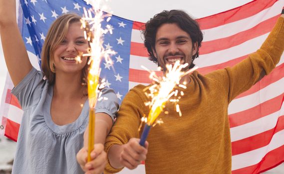 Portrait of young Caucasian couple playing with fire cracker while holding american flag. They are smiling and looking at camera