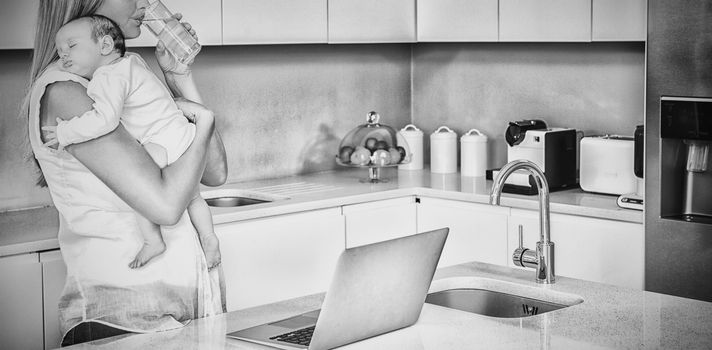 Young woman drinking water while carrying baby in kitchen at home