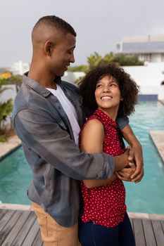 Front view of happy young mixed-race couple embracing each other and looking into each others eyes near swimming pool at home. 