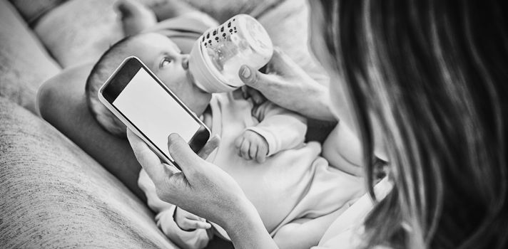 Mother using mobile phone while feeding her baby with milk bottle at home