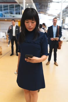 Close up of Asian businesswoman using mobile phone while standing in corridor at office with business people. International diverse corporate business partnership concept