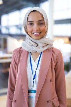 Portrait of beautiful smiling young mixed-race businesswoman in hijab with conference lanyard in a modern office. She is looking at camera. International diverse corporate business partnership concept