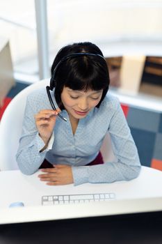 Overhead of pretty young Asian female customer service executive talking on headset at desk in office. This is a casual creative start-up business office for a diverse team