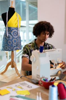 Front view of young mixed race male fashion designer using sewing machine on a table in design studio. This is a casual creative start-up business office for a diverse team