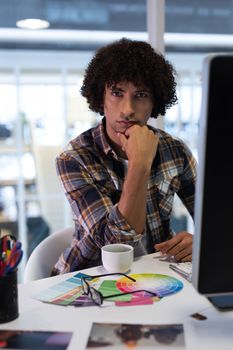 Portrait of young handsome mixed-race male graphic designer sitting hand on chin at desk in office. This is a casual creative start-up business office for a diverse team