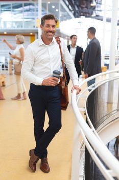 Front view of Happy caucasian businessman holding disposable coffee cup and looking at camera near railing in a modern office. International diverse corporate business partnership concept