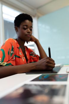 Side view of young pretty African american female graphic designer working on graphic tablet at desk in office. This is a casual creative start-up business office for a diverse team