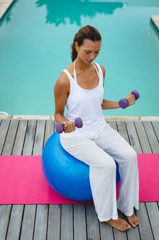 High angle view of fit mixed-race woman exercising with dumbbells while sitting on a exercise ball near swimming pool in the backyard. Summer fun at home by the swimming pool