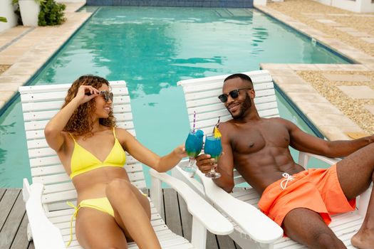 Front view of happy diverse couple toasting glasses of cocktail while relaxing on a sun lounger near swimming pool. Summer fun at home by the swimming pool