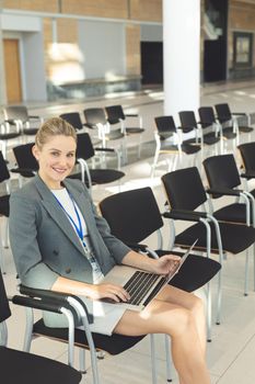 Side view of young businesswoman sat in empty conference room with laptop, smiling at camera