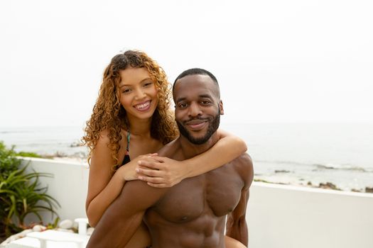 Portrait of African-american man giving piggyback ride to mixed-race woman near swimming pool at the backyard of home. Summer fun at home by the swimming pool