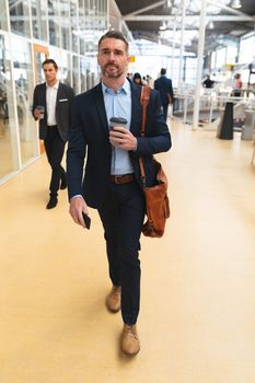 Front view of Caucasian businessman using mobile phone while walking in the corridor at office with business people. International diverse corporate business partnership concept
