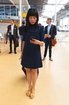 Front view of Asian businesswoman using mobile phone while standing in corridor at office with business people. International diverse corporate business partnership concept