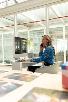 Side view of Caucasian female graphic designer talking on mobile phone while working on graphic tablet at desk. This is a casual creative start-up business office for a diverse team