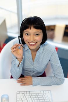 High angle view of pretty young Asian female customer service executive talking on headset at desk in office. This is a casual creative start-up business office for a diverse team