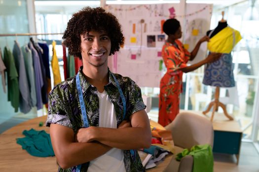Portrait of young mixed race male fashion designer sitting with arms crossed in design studio. African american woman working in the background. This is a casual creative start-up business office for a diverse team